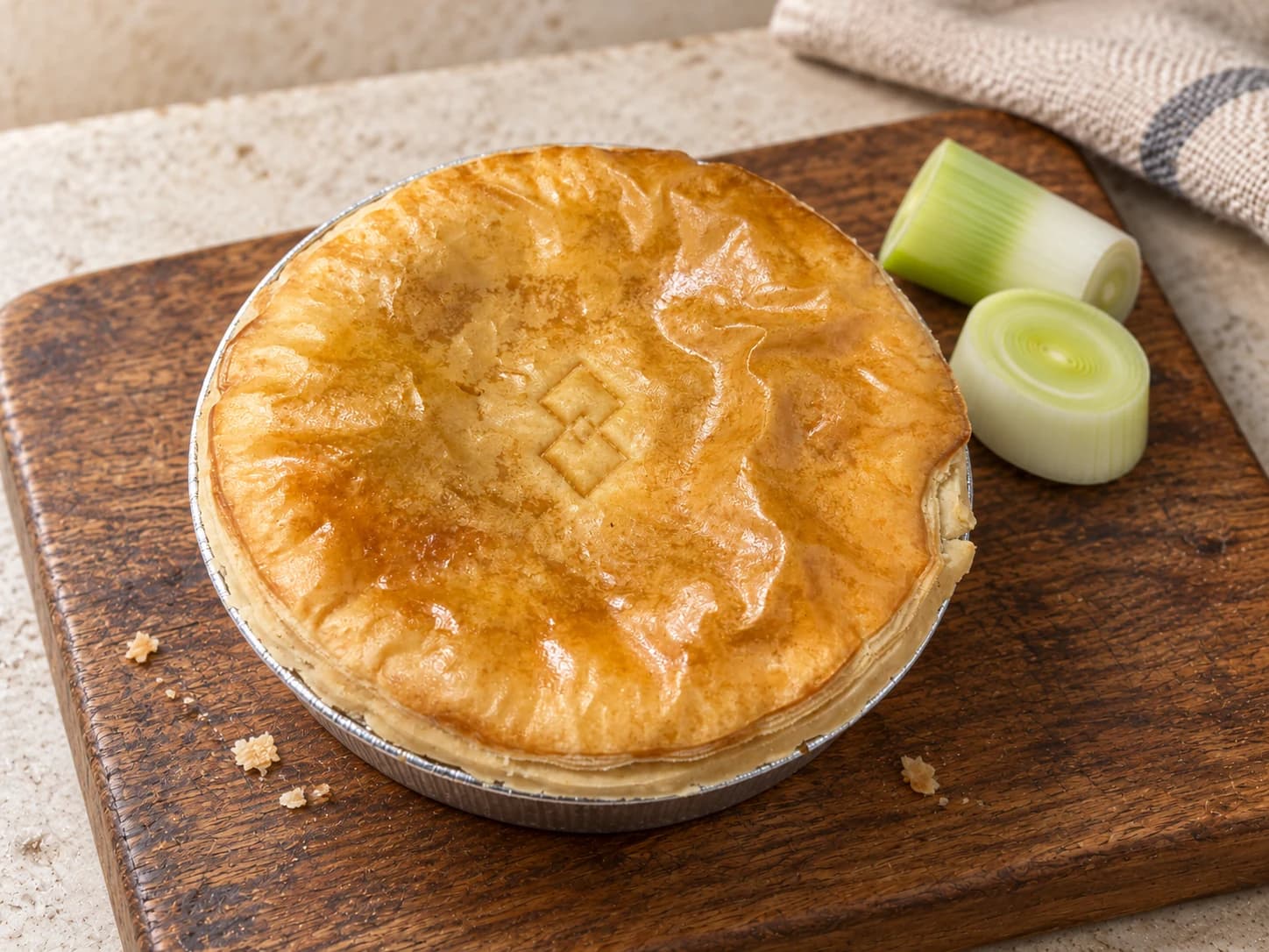 Pie with overlapping diamond impressions in its lid, beside pieces of leek on a wooden board.