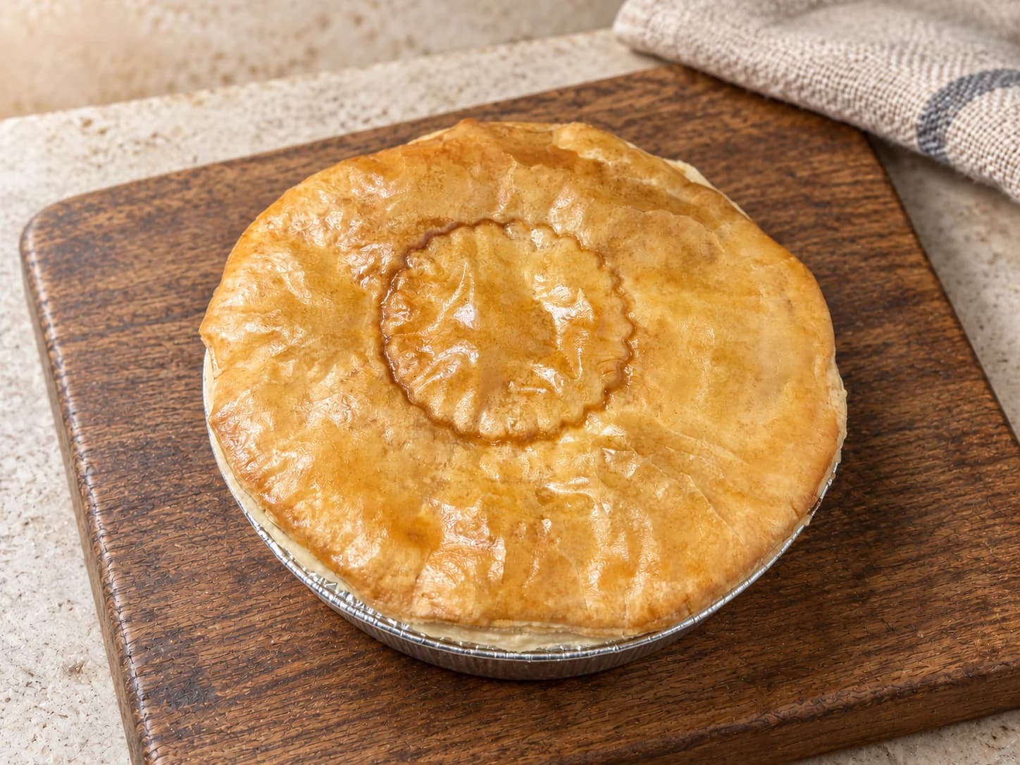 Pie with a small scalloped ring impression in its lid, in a foil case on a wooden board.