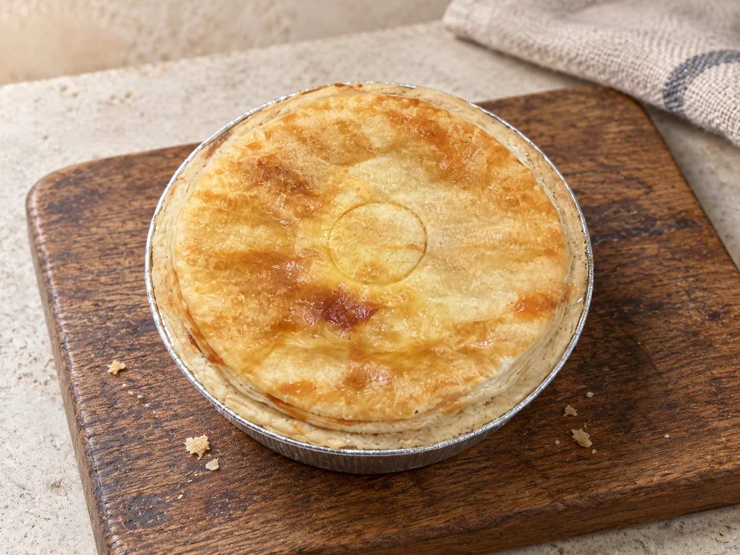 Golden pie with a small ring impression in its lid, in a foil case on a wooden board.