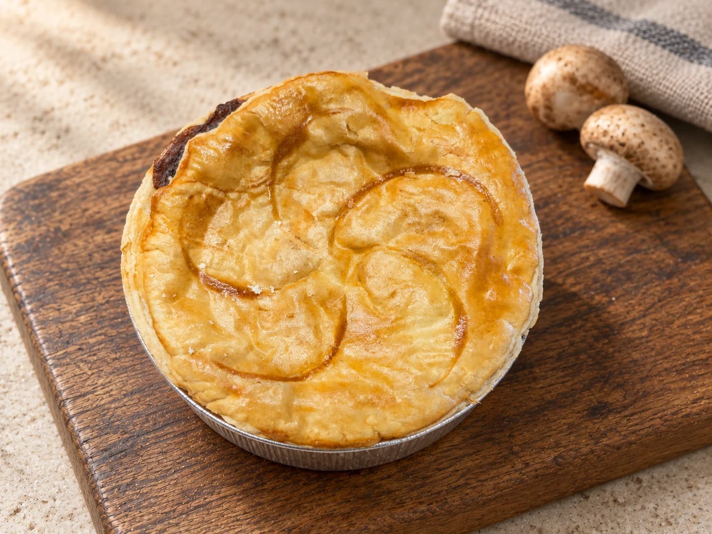 Pie with curved pinwheel scoring in its lid, in a foil case on a wooden board beside mushrooms.