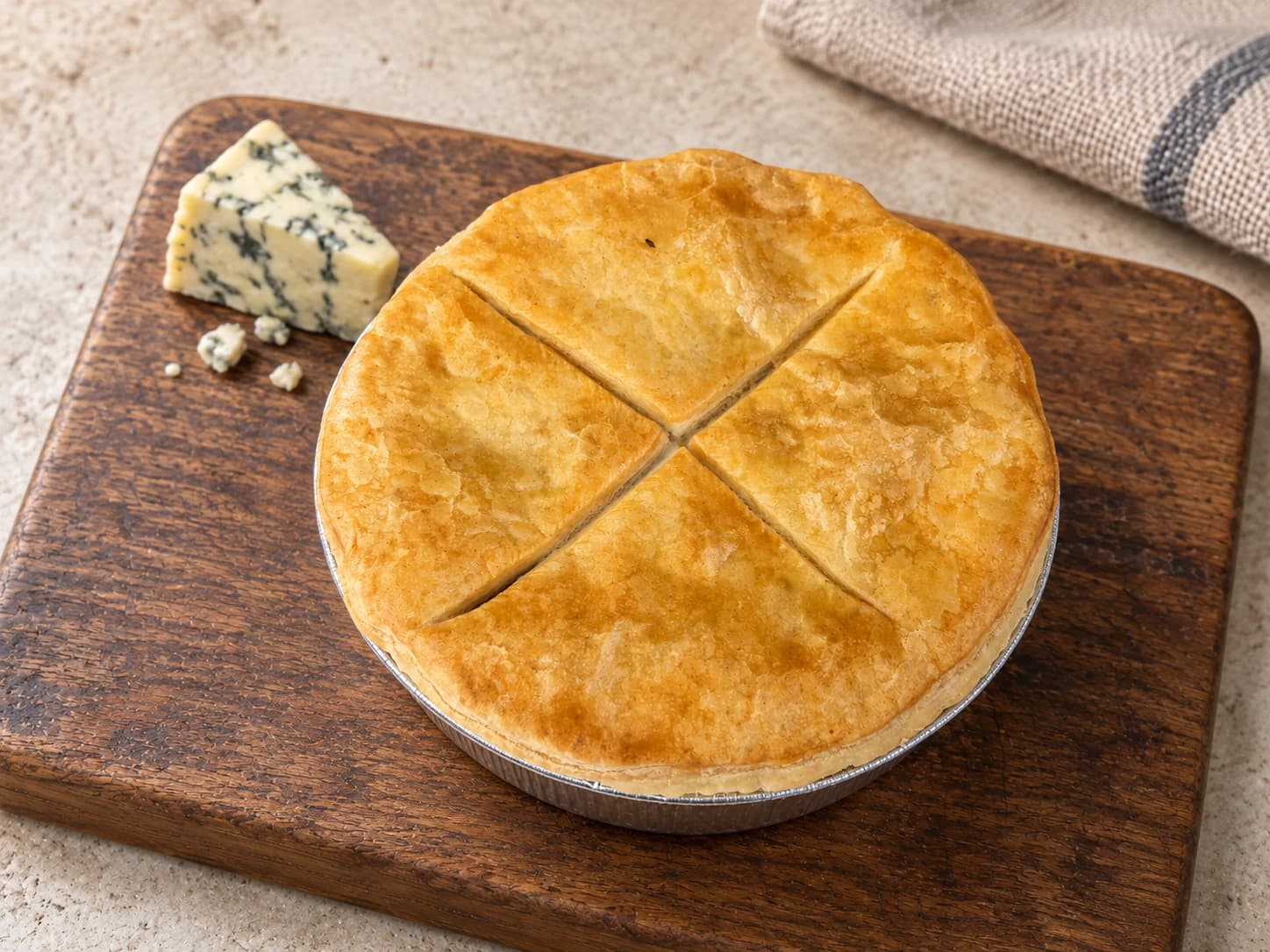 Pie with crossed scoring in its lid, in a foil case on a wooden board beside a wedge of blue cheese.