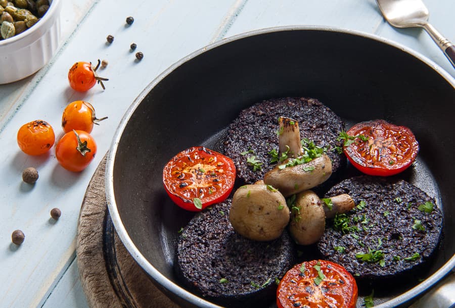 Black pudding slices with mushrooms and tomatoes in a frying pan.
