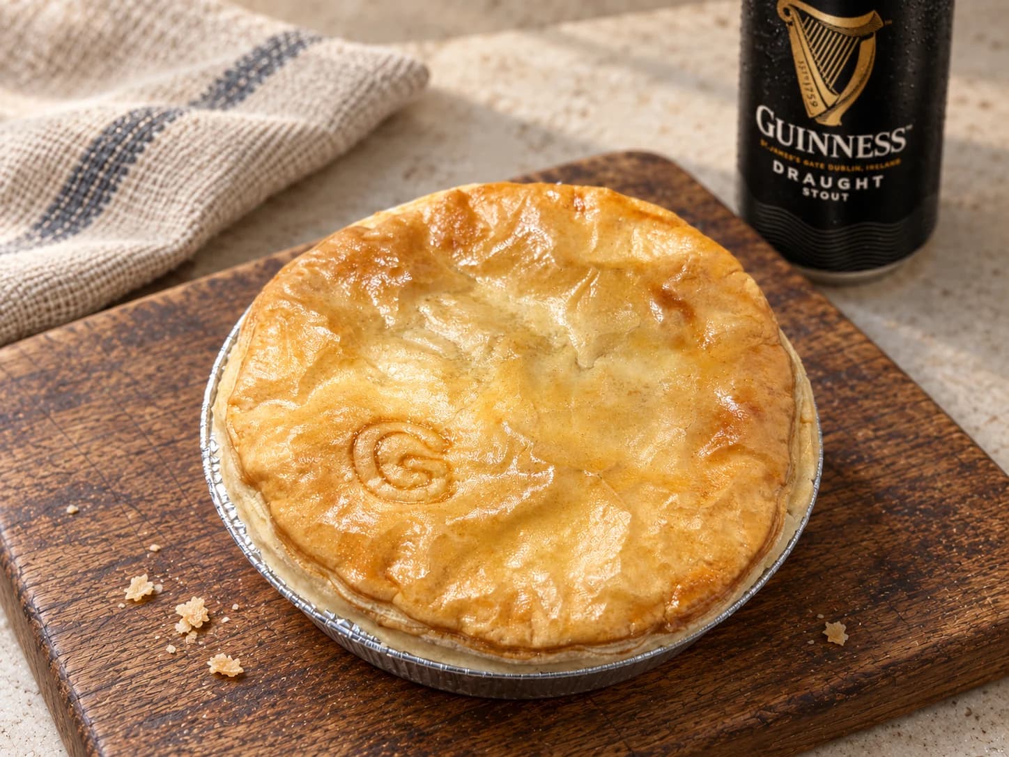 Pie with a small G-shaped impression, in a foil case on a wooden board with a Guinness can behind.